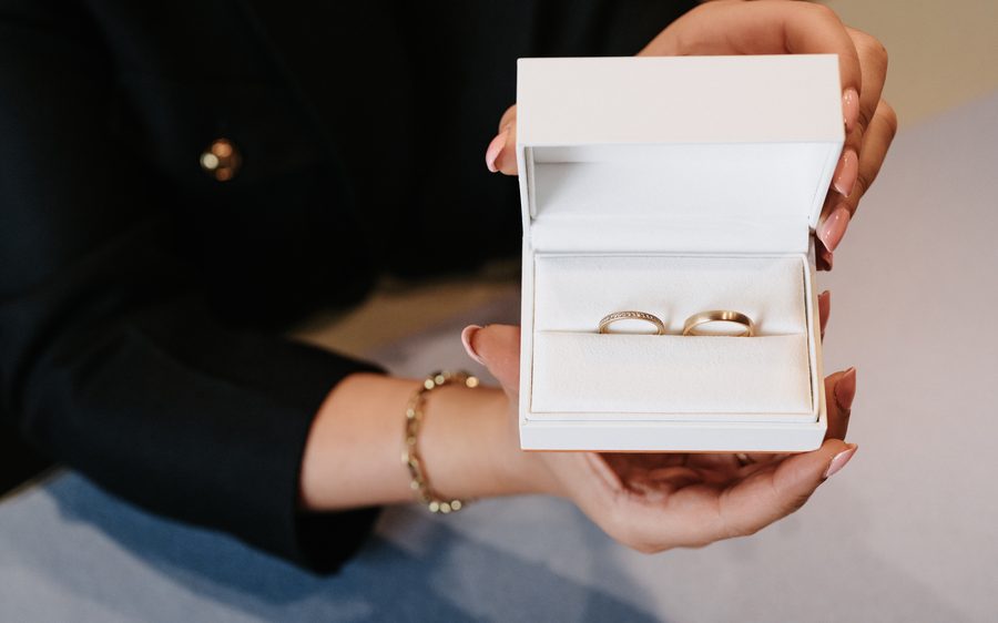 Zwei Hände halten eine geöffnete weiße Schachtel mit einem Trauringpaar in Gelbgold, ein Ring mit Brillantreihe