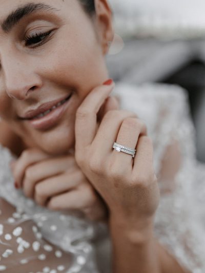 Braut lächelt und legt die Hand ans Kinn, am Ringfinger zwei schmale Ringe mit Brillanten